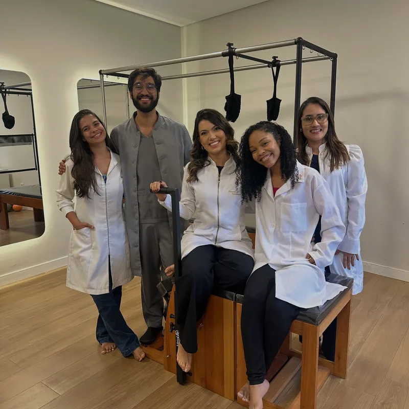 Cinco fisioterapeutas da CSI sorrindo na sala de Pilates, ao lado do aparelho de madeira, com piso amadeirado e espelho ao fundo.