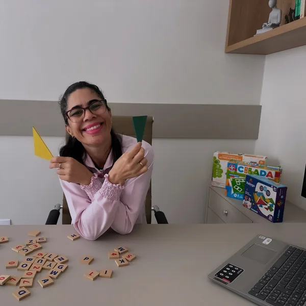 Psicopedagoga sorrindo à mesa de atendimento, com peças coloridas, letras de madeira e jogos pedagógicos.